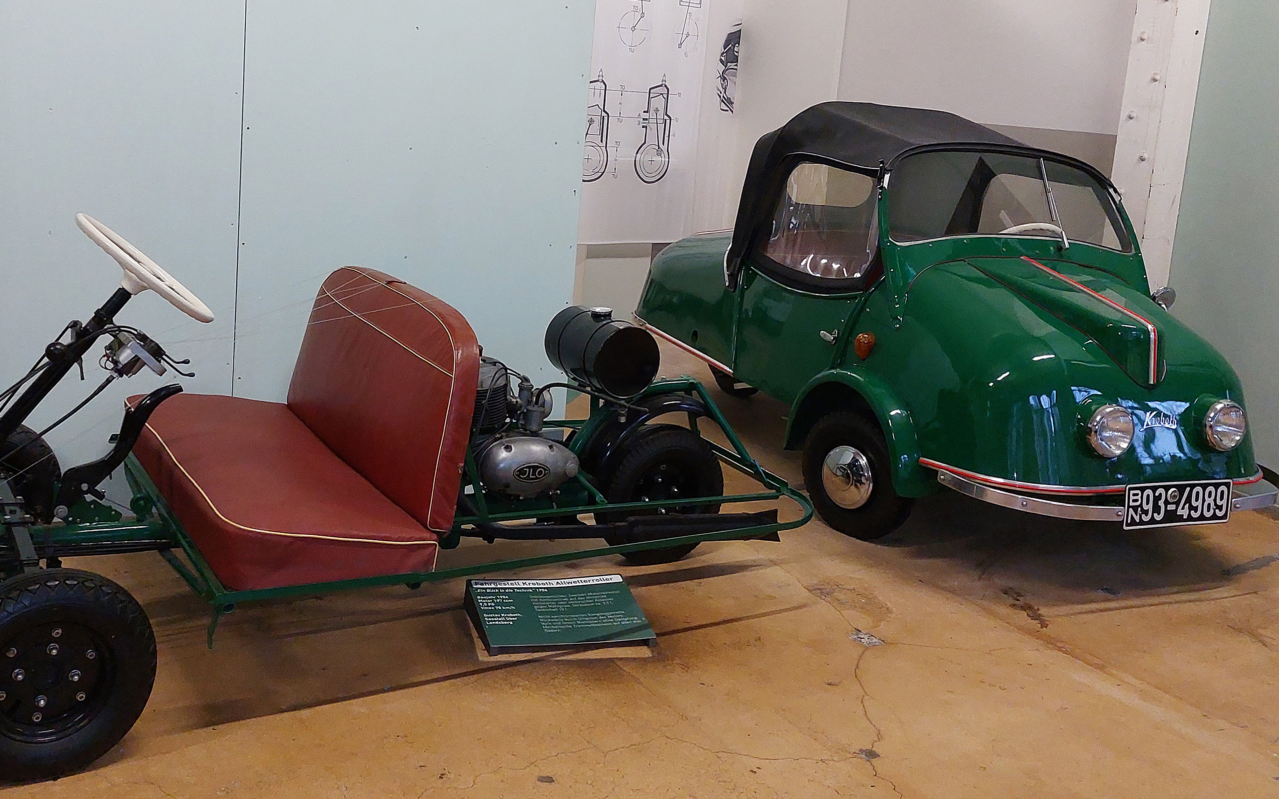 Auf dem Bild sind zwei Fahrzeuge zu sehen: Auf der linken Seite ein offenes, grünes Kart mit zwei Sitzen und einem Motor, rechts ein kleiner, geschlossener, grüner Wagen mit schwarzem Verdeck. Beide Fahrzeuge sind Teil einer Ausstellung über historische Autos.