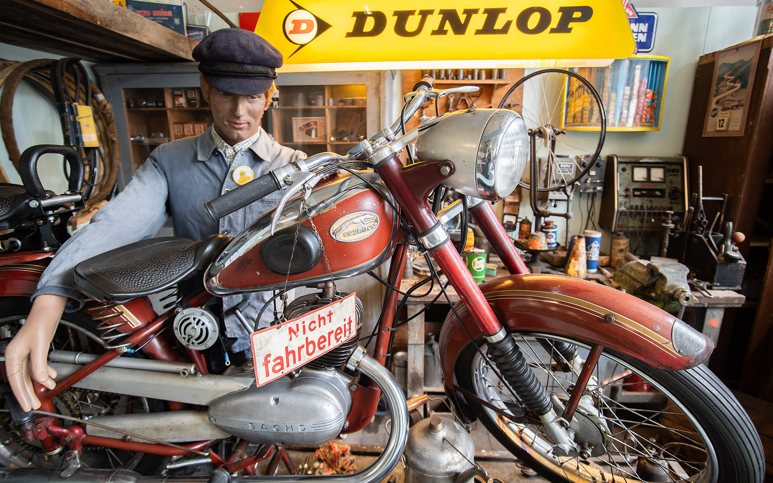 Das Bild zeigt ein altes Motorrad in einer Werkstatt, neben dem eine lebensgroße Statue eines Mechanikers steht. Am Motorrad hängt ein Schild mit der Aufschrift "Nicht fahrbereit". Im Hintergrund sind verschiedene Werkzeug- und Ersatzteilkisten sichtbar.