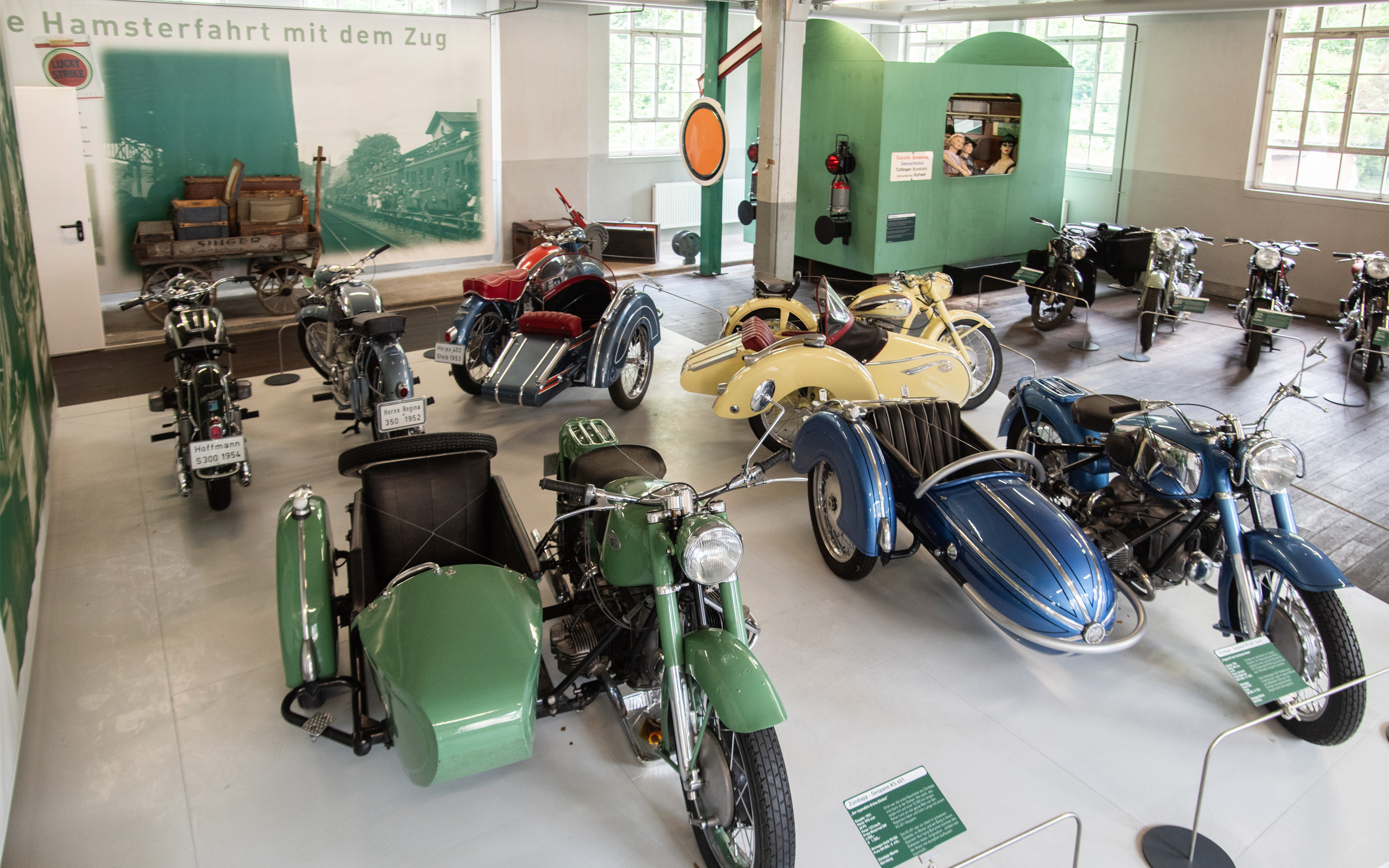 In einem Museum sind mehrere nostalgische Motorräder mit Beiwagen ausgestellt. Die Fahrzeuge in verschiedenen Farben sind ordentlich angeordnet, während im Hintergrund eine Wand mit der Aufschrift „Die Hamsterfahrt mit dem Zug“ zu sehen ist.