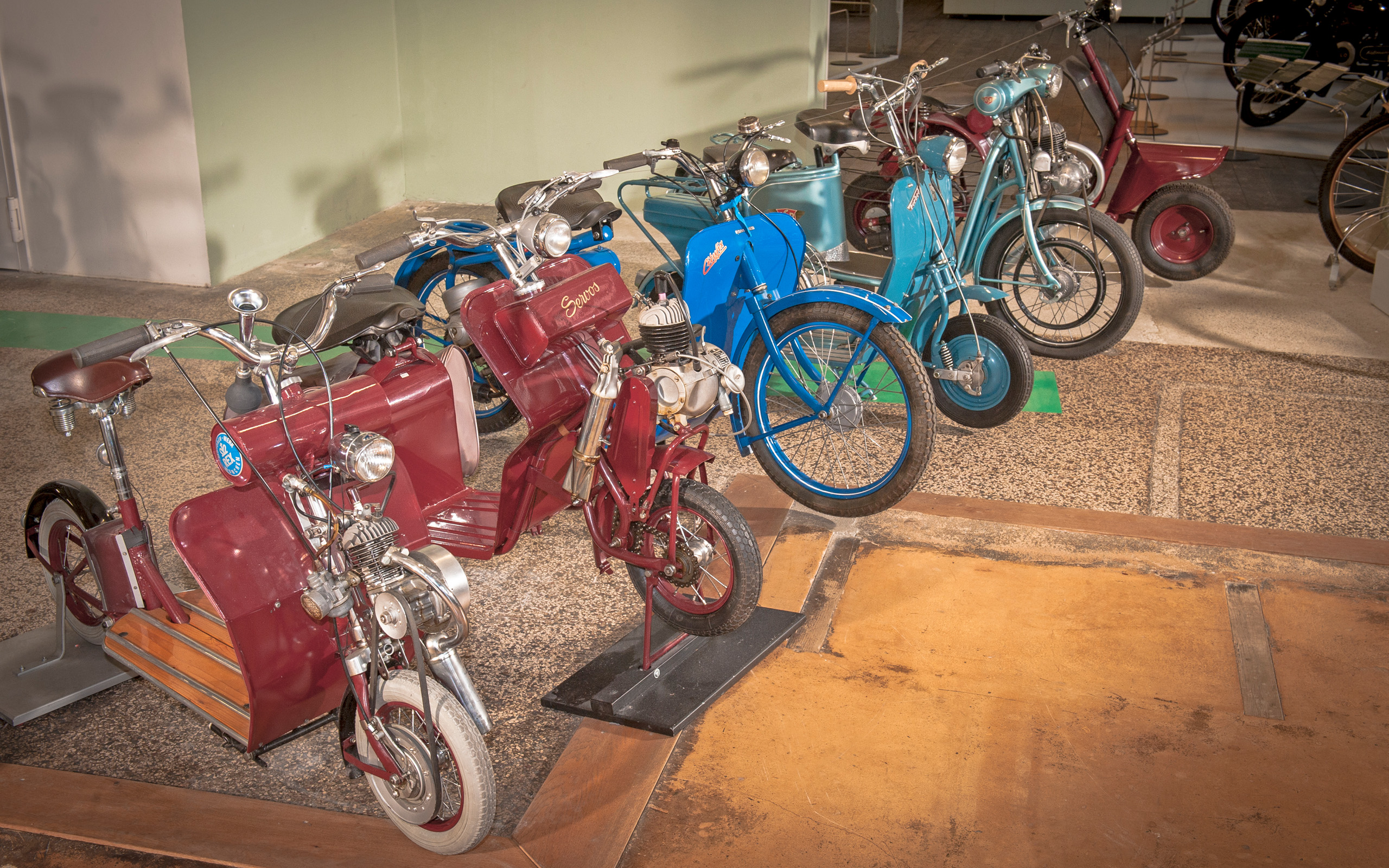 Eine Reihe von historischen Mopeds in verschiedenen Farben steht nebeneinander. Die Modelle zeigen ein nostalgisches Design, mit Holz- und Metallteilen. Ihre unterschiedlichen Farben, darunter Rot und Blau, verleihen der Ansammlung einen lebhaften Charakter. Die Mopeds reflektieren die Entwicklung der Zweiradtechnik.