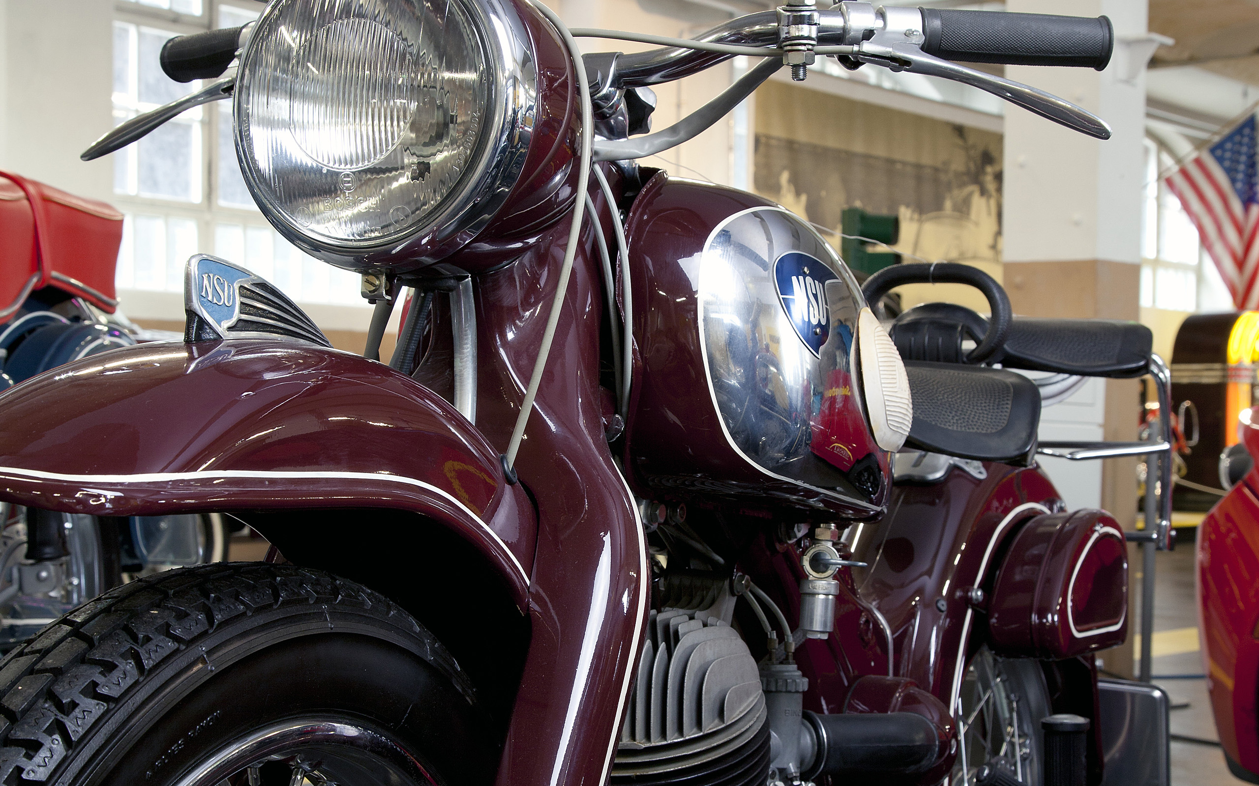 Ein rotes NSU-Motorrad, das im Vordergrund abgebildet ist. Die Scheinwerfer und das glänzende Logo sind gut sichtbar. Im Hintergrund sind weitere Motorradfahrzeuge und eine amerikanische Flagge zu erkennen. Das Bild vermittelt eine nostalgische Atmosphäre eines Motorradmuseums.