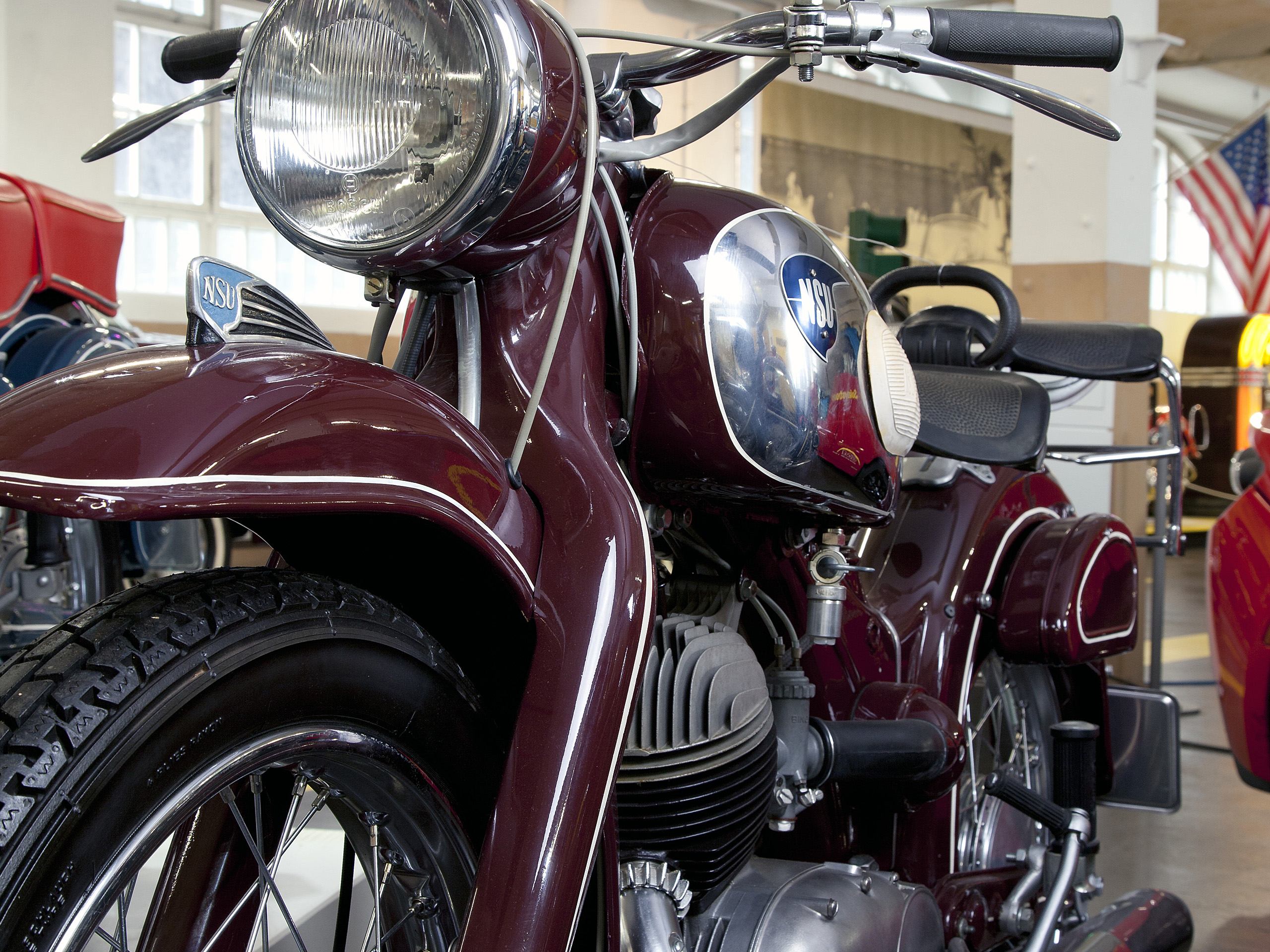 Ein klassisches Motorrad von NSU in dunklem Rot. An der Front ist der große Scheinwerfer gut sichtbar, und das Motorrad hat einen glänzenden Tank mit dem NSU-Logo. Die Umgebung wirkt wie ein Ausstellungsraum mit weiteren Fahrzeugen im Hintergrund.