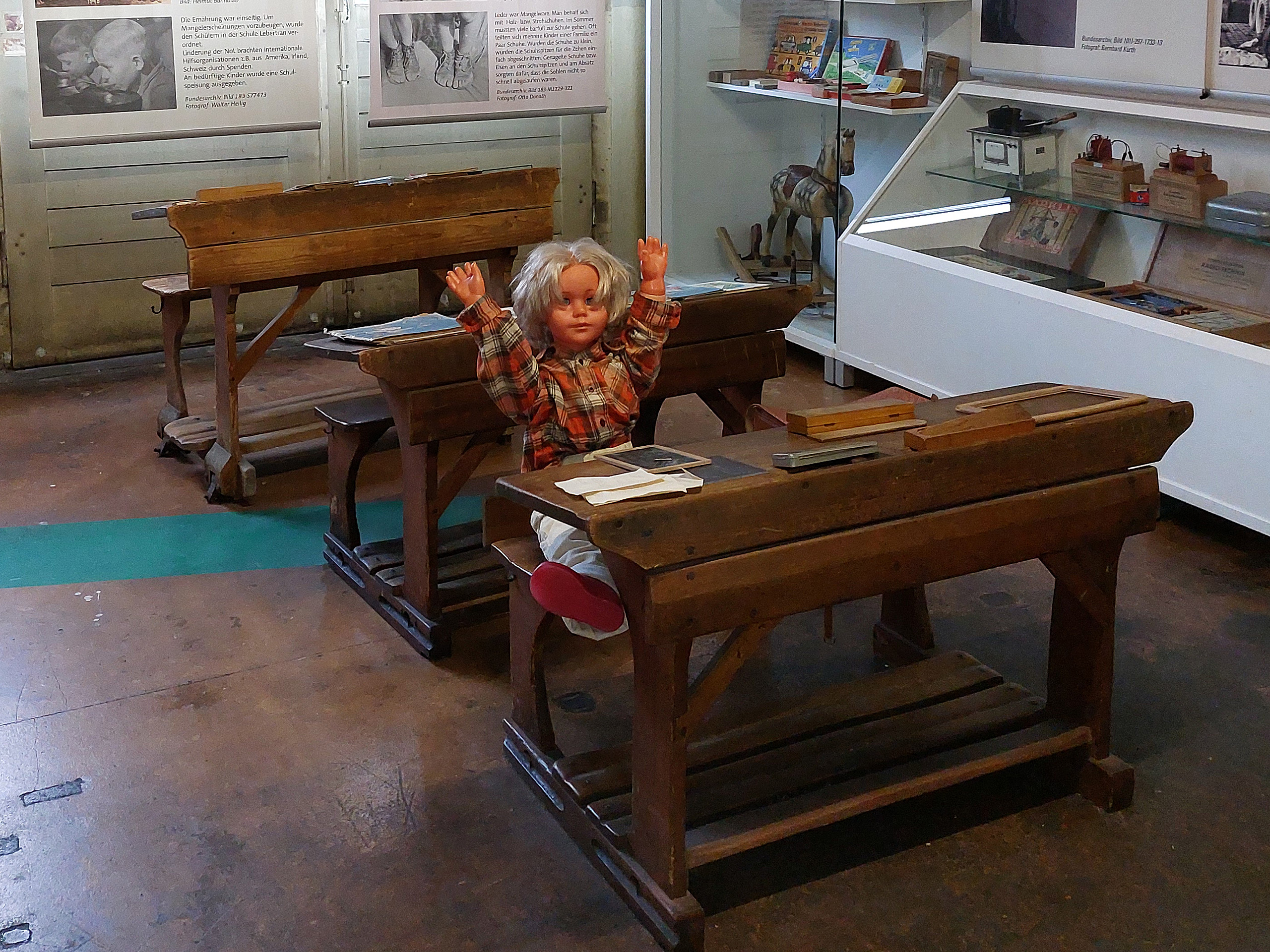 In einem Raum befinden sich zwei alte Schulbänke. Auf einer Bank sitzt eine Puppe mit blonden Haaren und einem karierten Hemd, die glücklich die Hände in die Höhe hebt. Der Boden ist aus Holz und es gibt eine Vitrine mit historischen Objekten im Hintergrund.