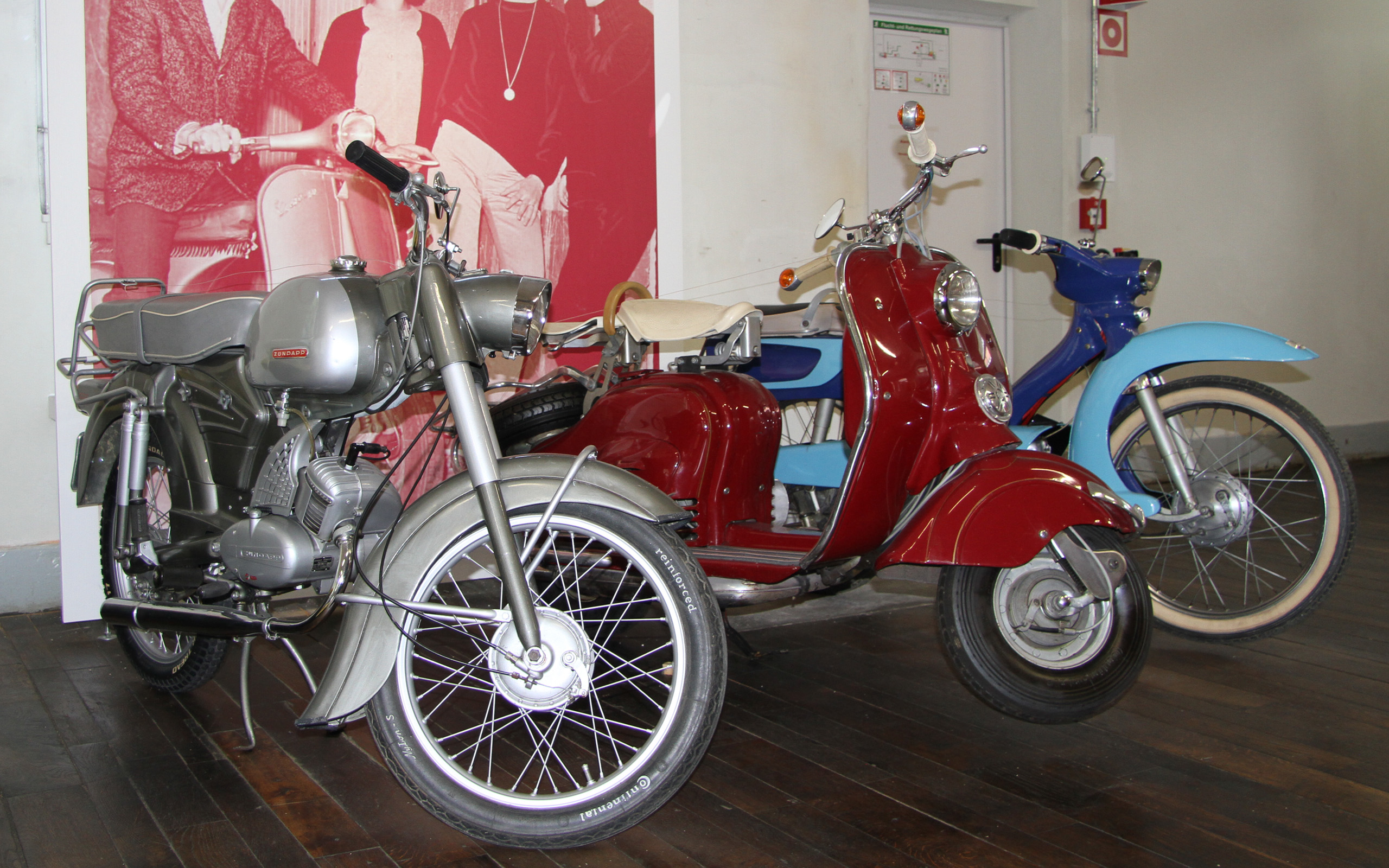 Drei alte Roller stehen in einem Raum: Ein silberner Roller auf der linken Seite, ein roter Roller in der Mitte und ein blauer Roller rechts. Der Fußboden ist aus Holz, und im Hintergrund sind unscharfe graue Wände zu sehen.