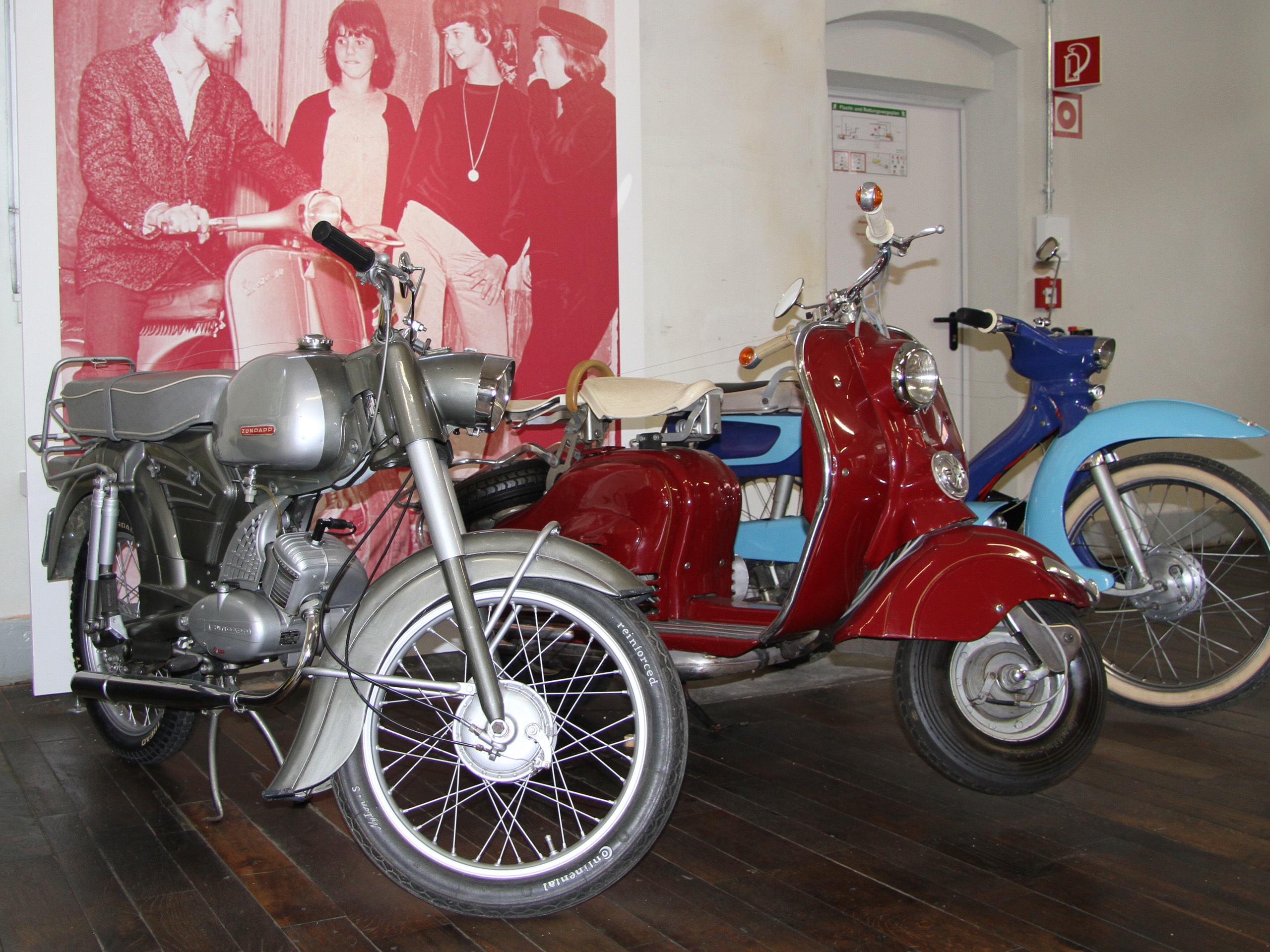 Das Bild zeigt zwei vintage Motorräder: links ein silbernes Fahrzeug mit einem schmalen Design und rechts ein rotes Scooter-Modell. Im Hintergrund sind verschwommene Figuren zu sehen, die in schwarz-weiß gehalten sind. Der Boden ist aus Holz und der Raum wirkt nostalgisch.