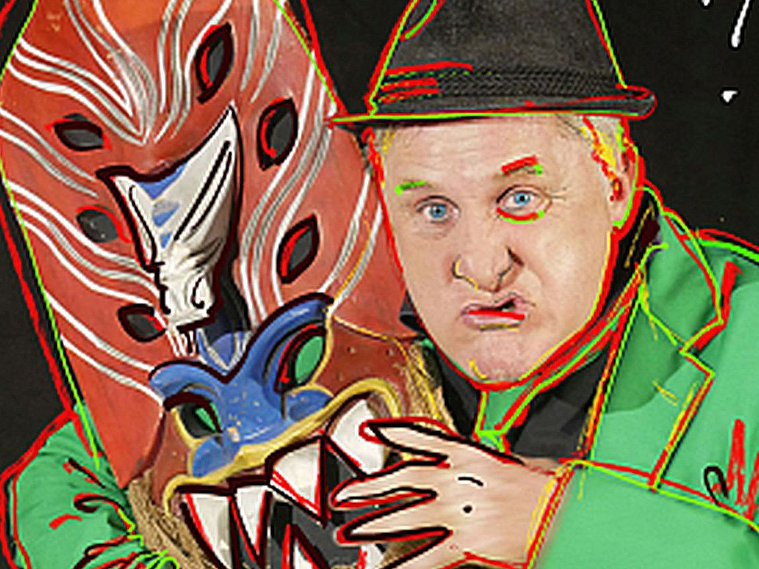 Ein Mann mit blonden Haaren trägt einen schwarzen Hut und eine grüne Jacke. Er hält eine farbenfrohe maskenähnliche Skulptur, die aussieht wie ein bunter Kopf mit Zähnen. Sein Gesichtsausdruck ist lustig und herausfordernd, ideal für eine unterhaltsame Atmosphäre.