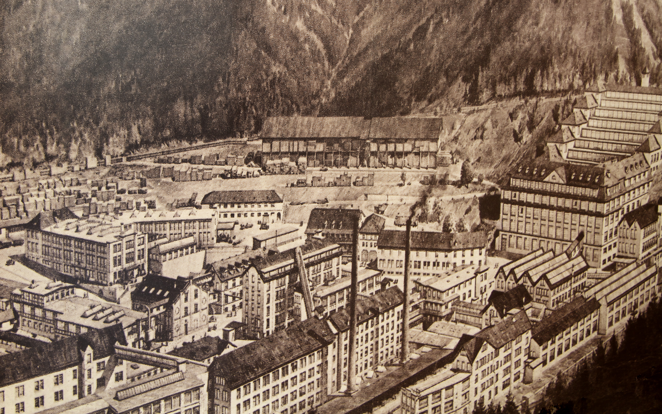 Die Aufnahme zeigt eine historische Fabriklandschaft in einem gebirgigen Gebiet. Verschiedene Gebäude, darunter Fabriken und Lagerhäuser, sind zu sehen. Im Hintergrund erheben sich hohe Berge, die die Kulisse bilden. Die gesamte Szene vermittelt ein Gefühl industrieller Aktivität und Entwicklung.