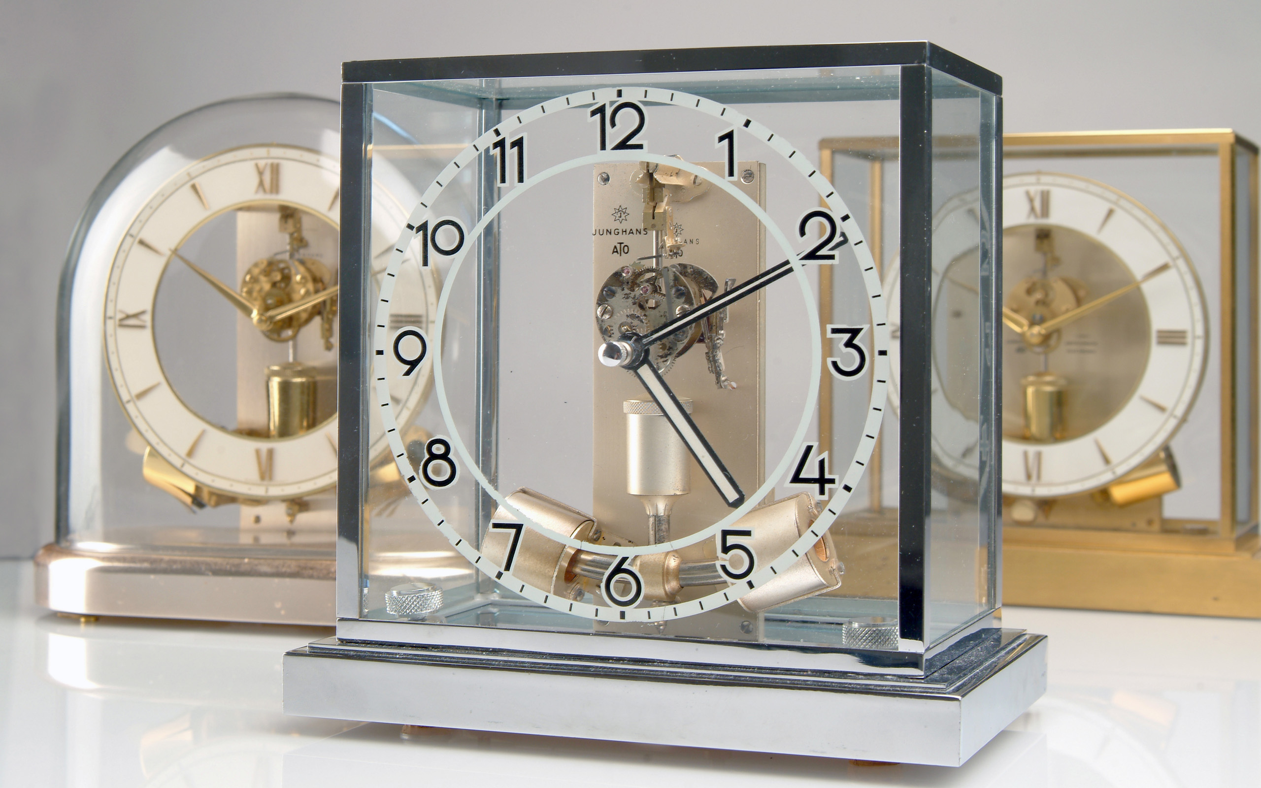 Eine moderne ATO Tischuhr mit klaren Linien und einem transparenten Gehäuse. Der Ziffernblatt zeigt große schwarze Zahlen und zwei Zeiger, die die aktuelle Zeit anzeigen. Im Hintergrund sind zwei weitere Uhren sichtbar, die unterschiedliche Designs haben.