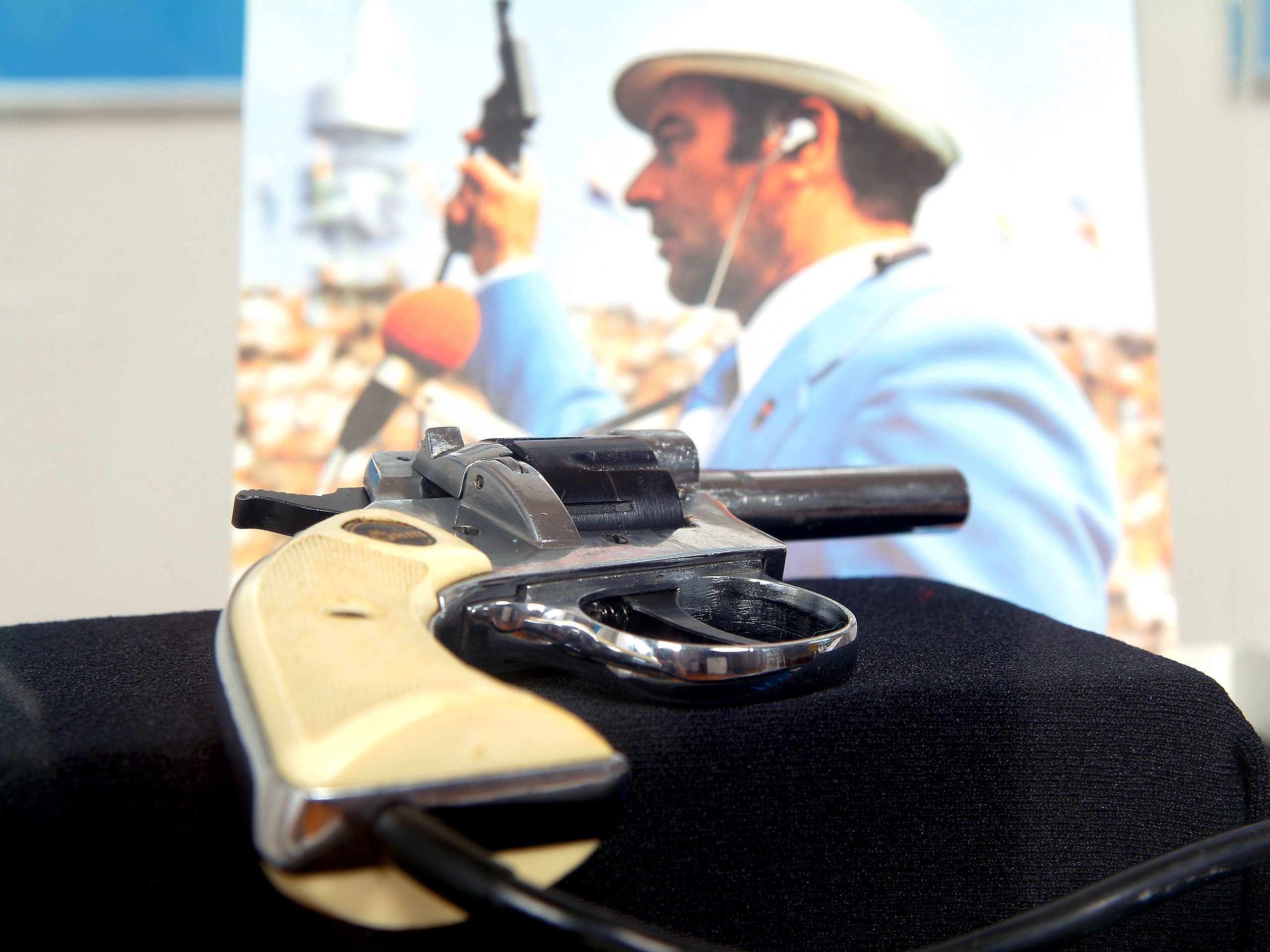 Ein leicht diagonal positionierte Startpistole wird im Vordergrund gezeigt, während im Hintergrund eine Person bei einer Sportveranstaltung mit Startpistole, die einen Anzug trägt, zu sehen ist. 