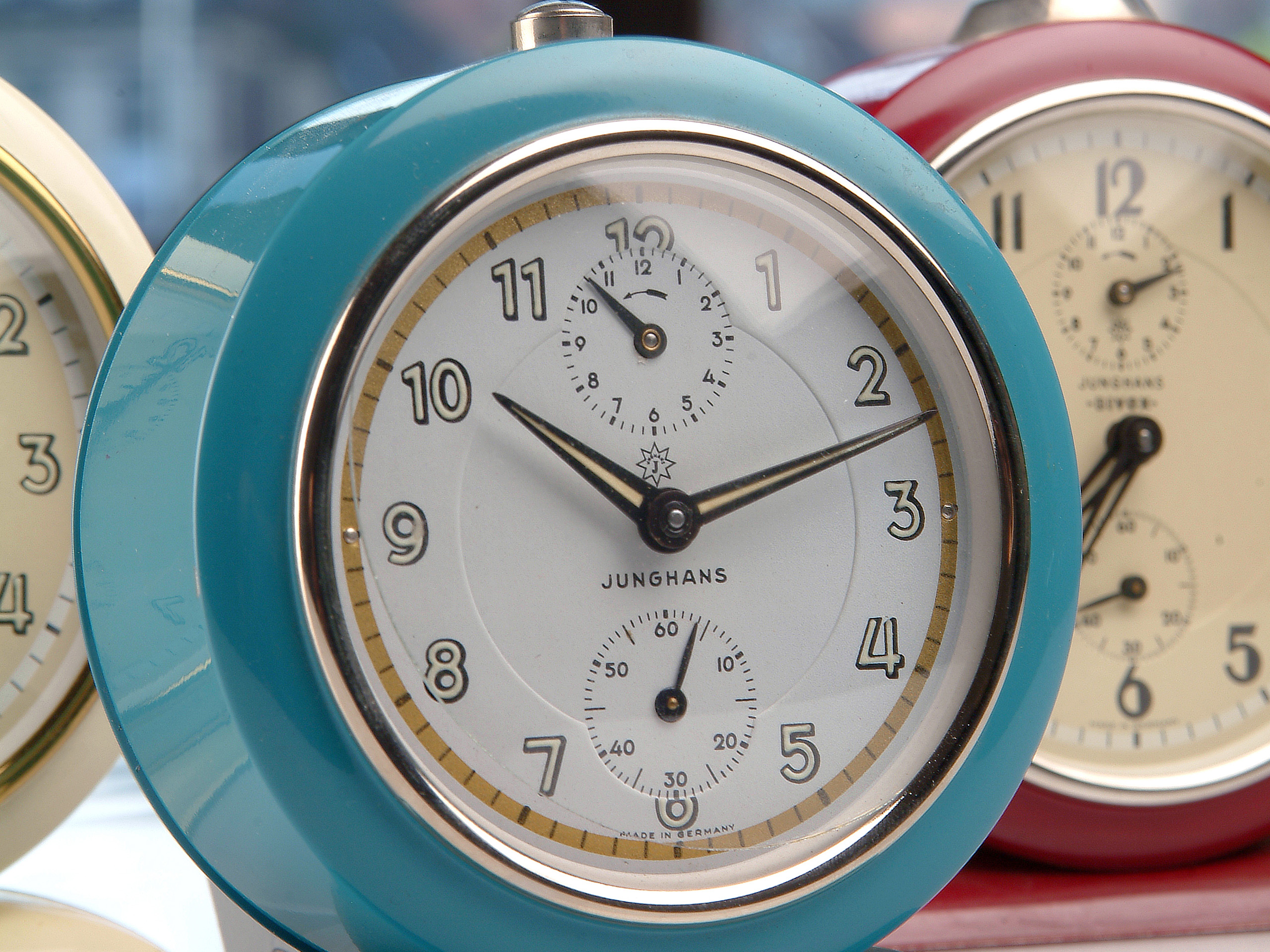 Drei Wecker in verschiedenen Farben stehen nebeneinander. Der vorderste ist in einem leuchtenden Türkis gehalten und hat römische Ziffern auf dem Ziffernblatt. Es gibt zusätzlich einen kleinen Sekundenzeiger. Die Marke „Junghans“ ist sichtbar.