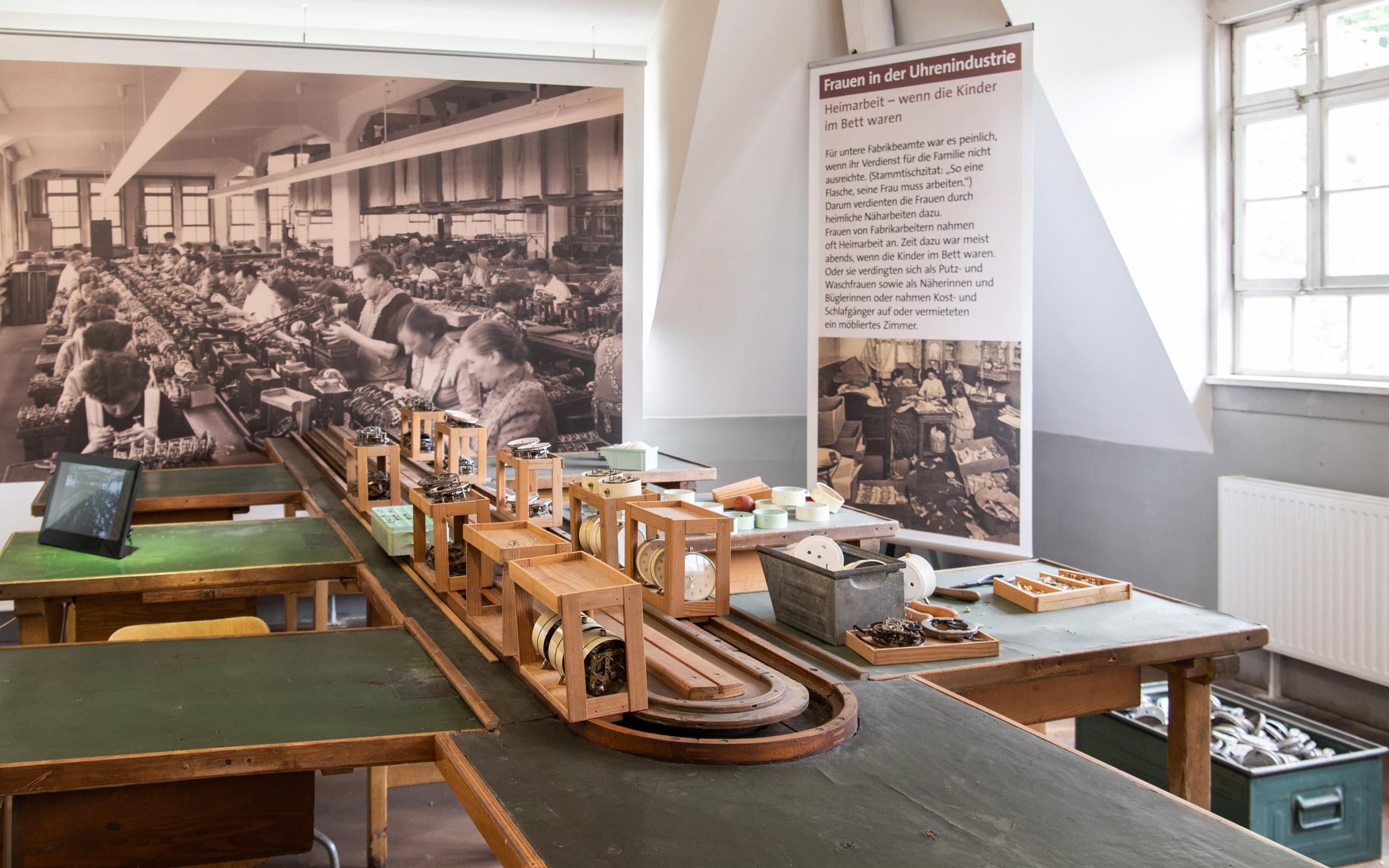Das Bild zeigt einen Arbeitsraum mit Holztischen, auf denen verschiedene Uhrenbestandteile liegen. Im Hintergrund ist ein historisches Foto von Frauen, die Uhren montieren. Ein Informationsbanner thematisiert die Rolle von Frauen in der Uhrenindustrie und deren Herausforderungen in der Heimarbeit.