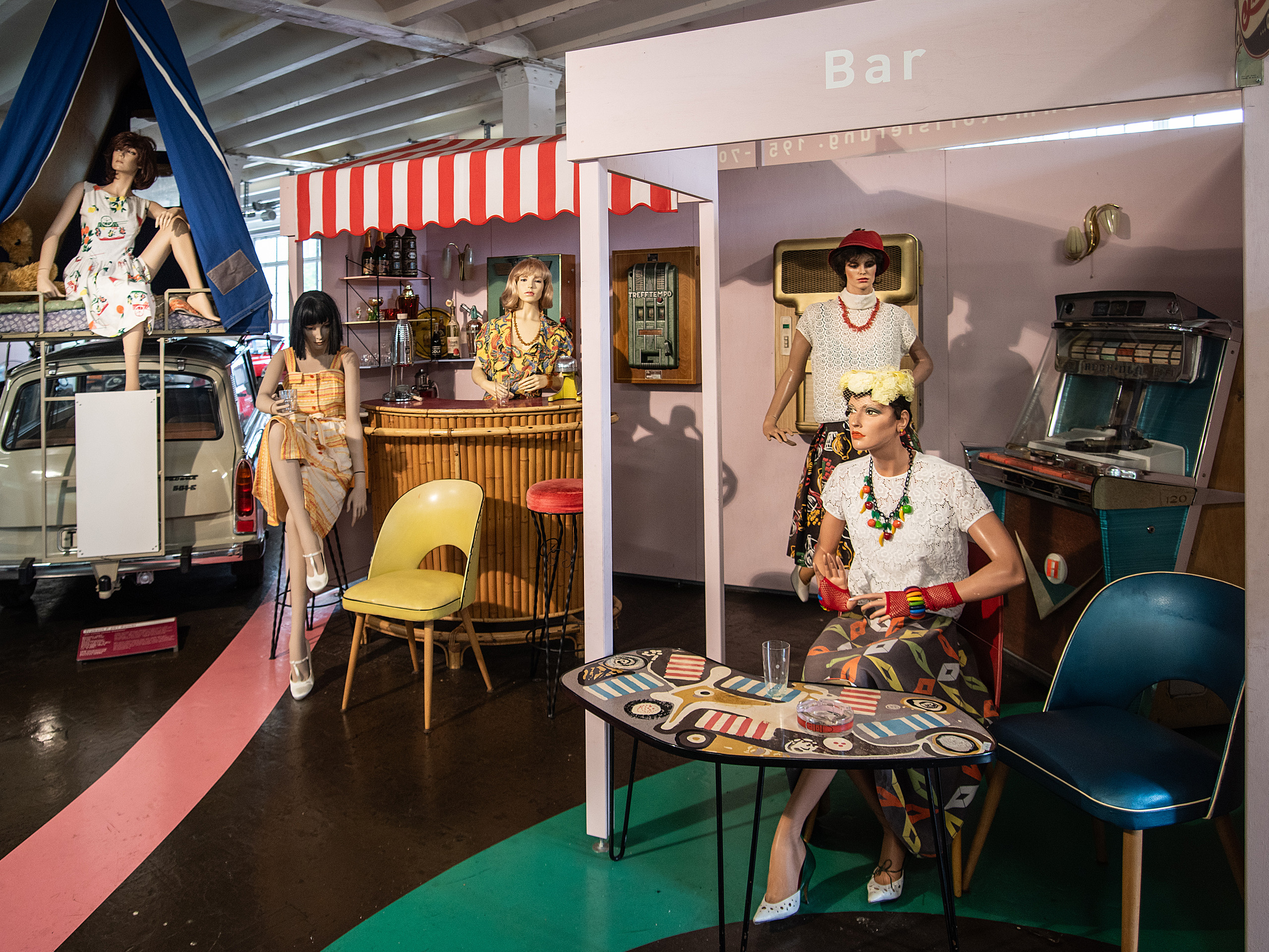 Die Szene zeigt eine Retro-Bar mit mehreren Schaufensterpuppen in verschiedenen ModeOutfits. Eine Puppe sitzt an einem Tisch, während andere geduldig an der Bar stehen. Der Raum ist bunt gestaltet, mit einer nostalgischen Atmosphäre und typischen 60er-Jahre-Elementen.