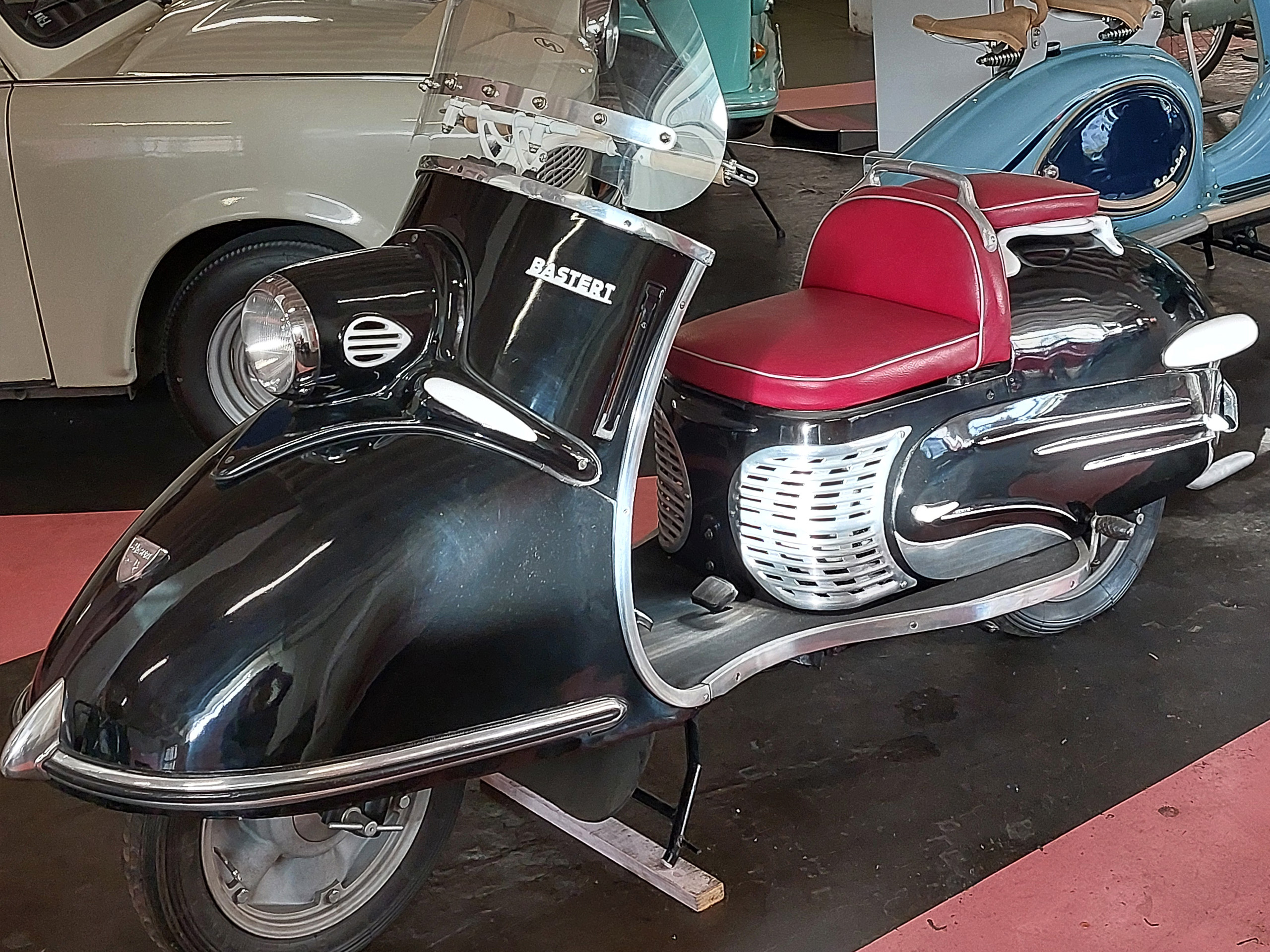 Ein klassischer Motorroller in Schwarz mit einem roten Sitzpolster. Der Roller hat abgerundete Linien und eine verchromte Verkleidung an der Seite. Im Hintergrund sind weitere Fahrzeuge zu sehen, die eine Retro-Atmosphäre vermitteln.