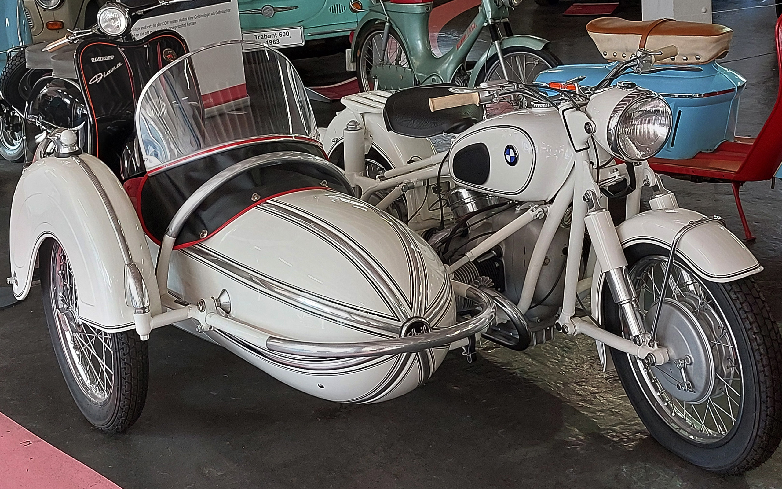 Das Bild zeigt ein klassisches, weißes Motorrad mit einem Beiwagen. Das Motorrad hat ein markantes Design und ist mit zahlreichen verchromten Elementen versehen. Der Beiwagen ist ebenfalls in Weiß gehalten und trägt dekorative Linien. Im Hintergrund sind weitere Oldtimer zu sehen.