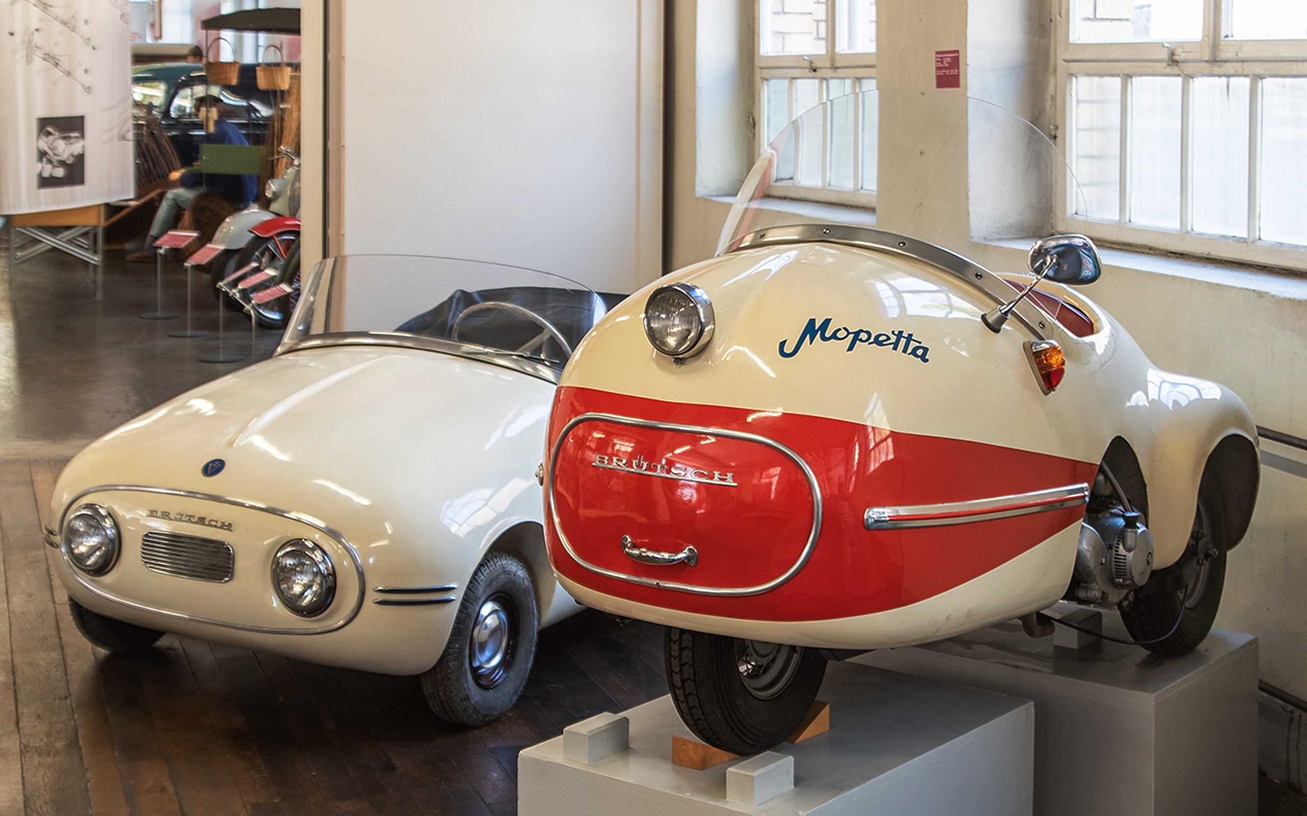 Das Bild zeigt zwei historische Fahrzeuge in einem Museum. Links befindet sich ein weißes, kompaktes Auto mit runden Scheinwerfern. Rechts steht ein dreirädriges Fahrzeug namens Mopetta, das eine rot-weiße Lackierung hat und ein markantes, futuristisches Design aufweist.