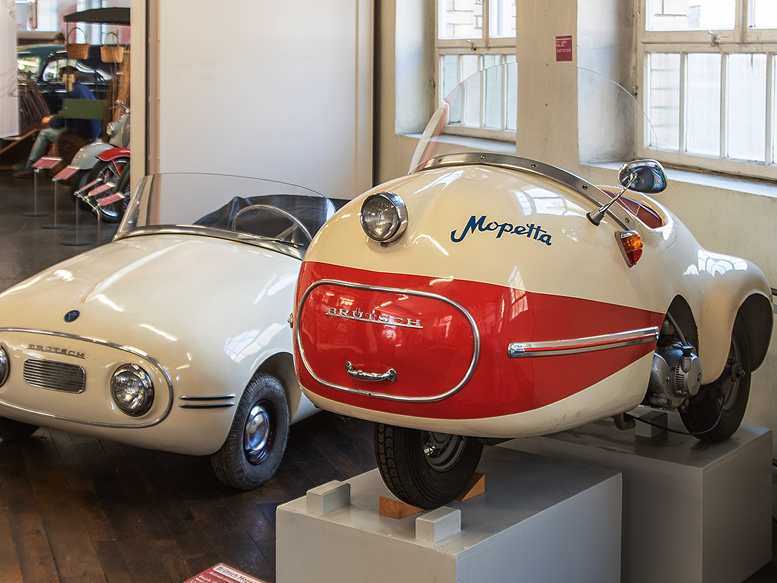 Das Bild zeigt zwei historische Fahrzeuge in einem Museum. Links befindet sich ein weißes, kompaktes Auto mit runden Scheinwerfern. Rechts steht ein dreirädriges Fahrzeug namens "Mopetta", das eine rot-weiße Lackierung hat und ein markantes, futuristisches Design aufweist.