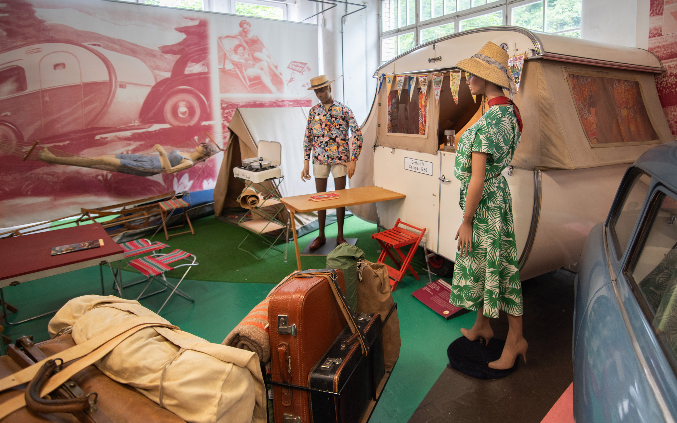 Ein Ausstellungsraum zeigt eine nostalgische Camping-Szene. Eine Puppe in einem grünen Kleid steht neben einem farbenfrohen Wohnwagen, während eine weitere Puppe in einem bunten Hemd steht. Gepäck und Campingmöbel sind sichtbar, der Boden ist mit grünem Teppich bedeckt.