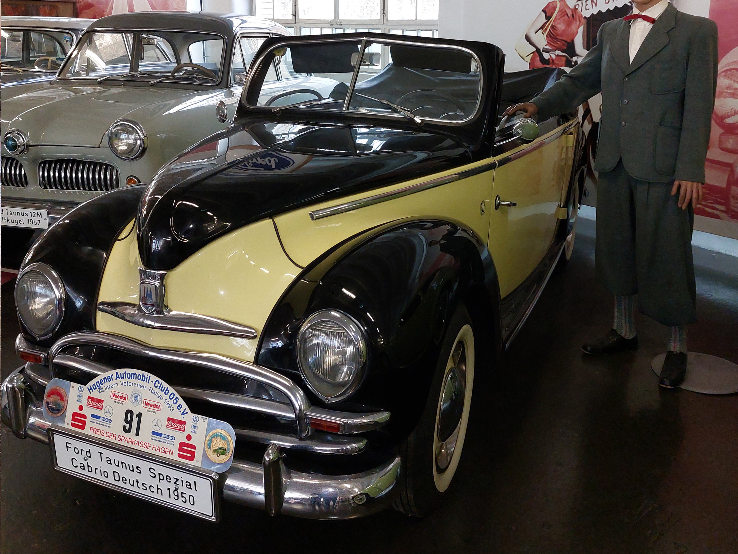 Ein schwarzes und gelbes Cabriolet, ein Ford Taunus Spezial von 1950, steht in einem Automuseum. Neben dem Auto steht eine figurative Darstellung eines Mannes in Anzug und Fliege. Im Hintergrund sind weitere Oldtimer zu sehen.