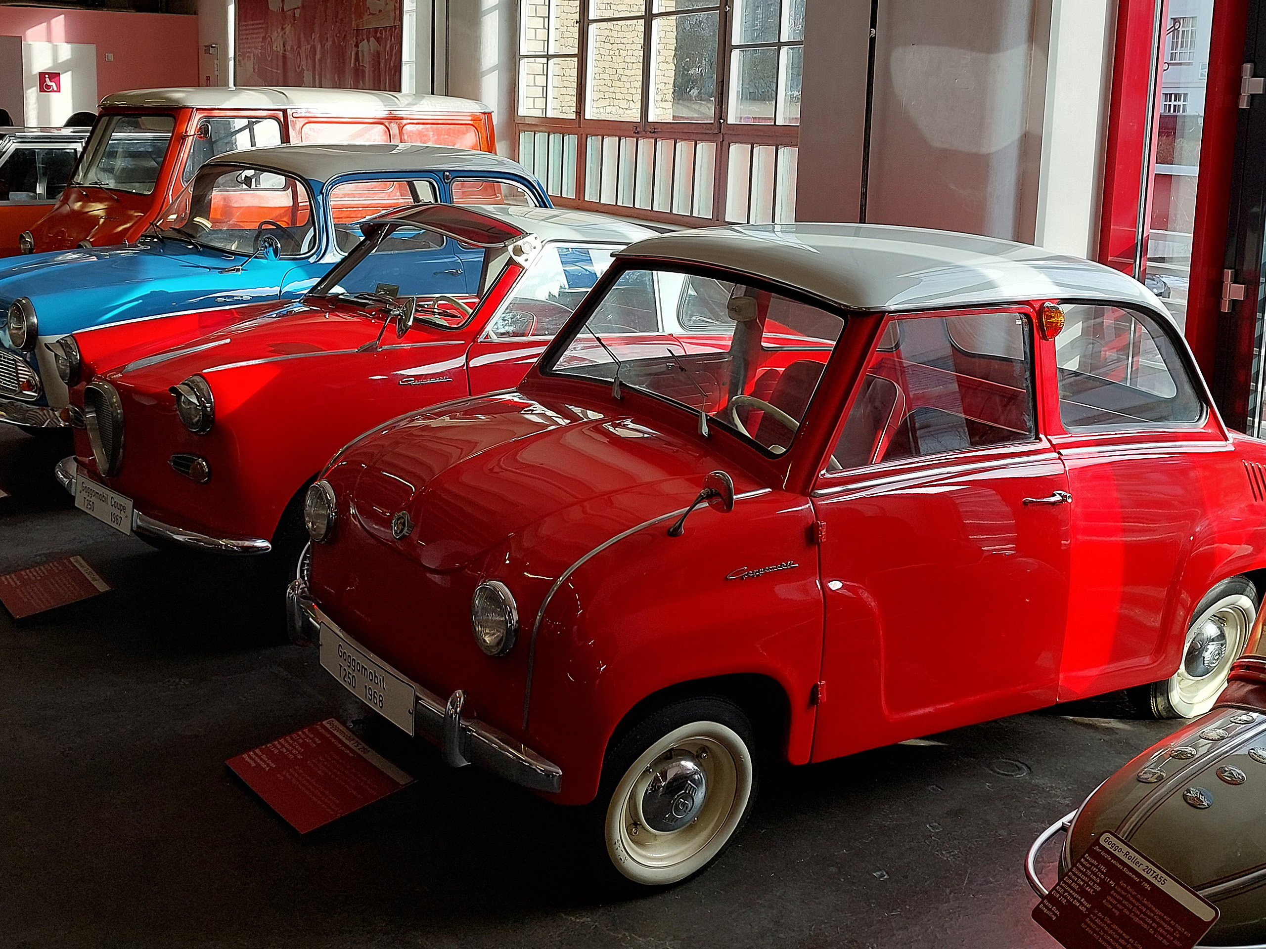Das Bild zeigt mehrere klassische Autos in einem Museum. Im Mittelpunkt steht ein rotes Auto mit einem grauen Dach, das einen nostalgischen Charme ausstrahlt. Daneben sind ein blauer und ein weiterer roter Wagen sichtbar, alles in gutem Zustand und umgeben von natürlichem Licht.
