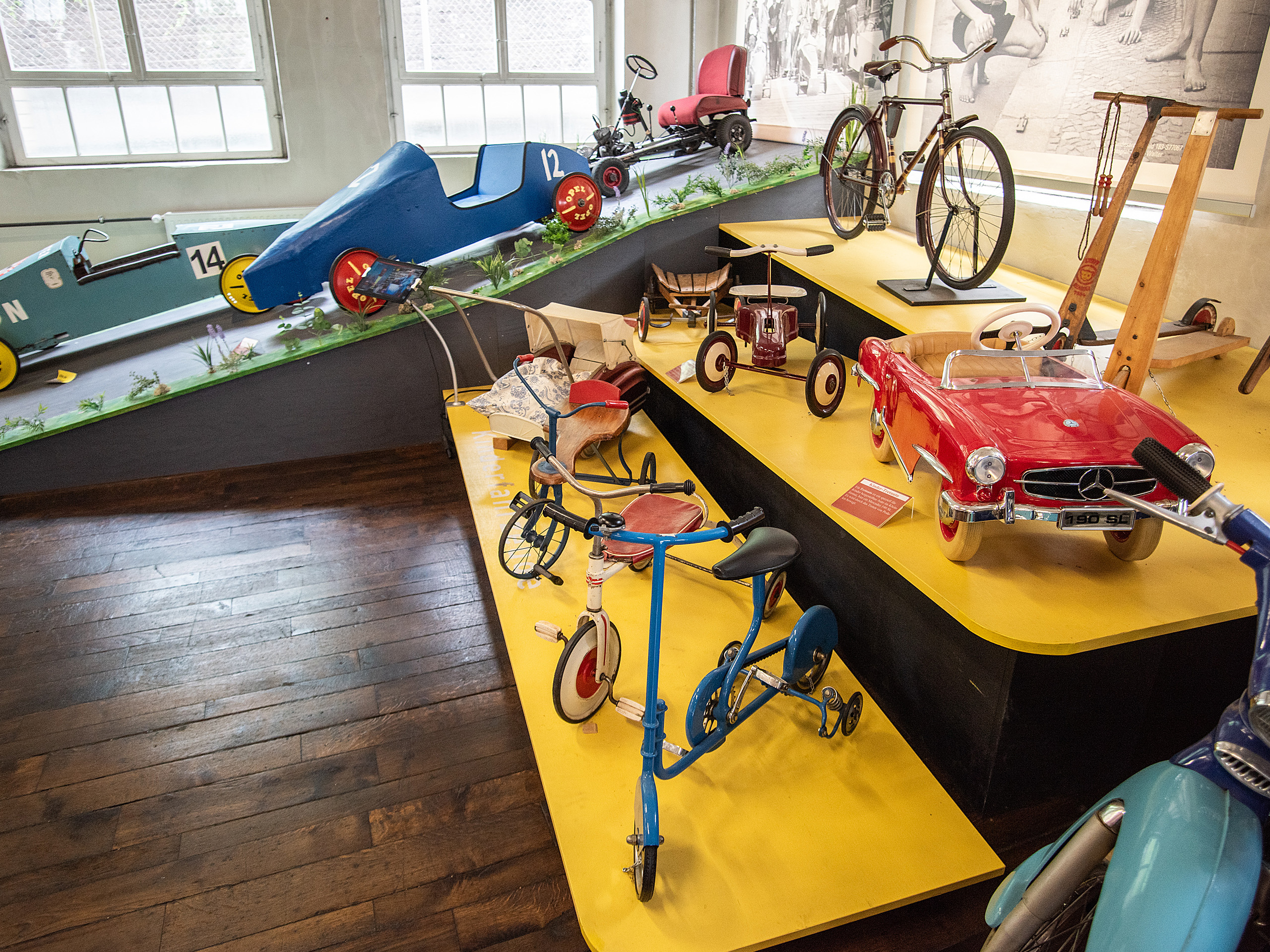 Im Bild ist eine Ausstellung mit vintage Spielzeugen zu sehen. Darunter befinden sich Holzfahrzeuge, darunter ein blauer Dreirad, ein rotes Spielauto und verschiedene Fahrräder. Der Boden ist aus Holz und die Plattformen sind gelb. Die Umgebung wirkt nostalgisch und einladend.