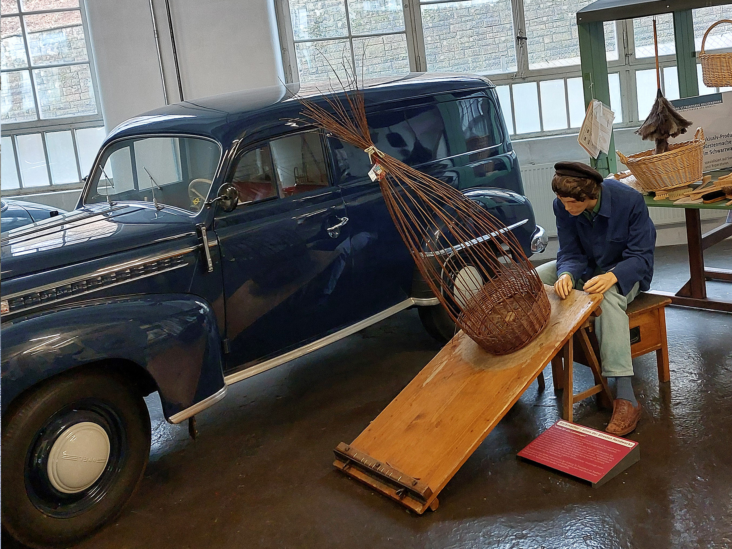 Eine Wachsfigur zeigt einen Handwerker, der an einem Tisch sitzt und an einem Weidenkorb arbeitet. Neben ihm steht ein schräger Tisch, auf dem der Korb platziert ist. Im Hintergrund ist ein blauer Oldtimer zu sehen. Das Bild vermittelt handwerkliche Tradition.