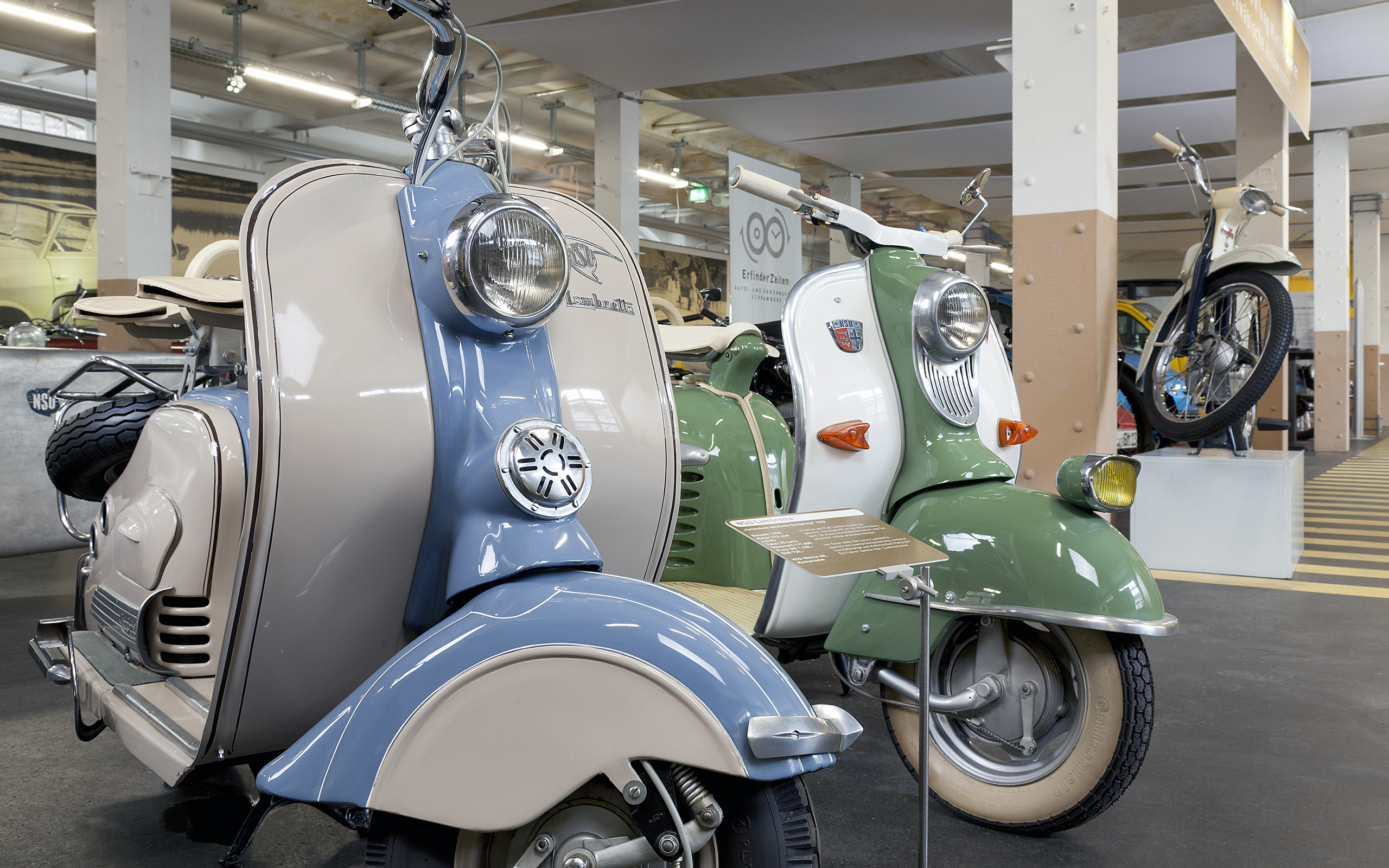 Zwei historische Scooter stehen in einem Museum ausgestellt. Der linke Scooter ist in sanften Creme- und Blautönen gehalten, während der rechte grün und weiß ist. Beide Fahrzeuge haben runde Scheinwerfer und nostalgisches Design, das Retro-Charme ausstrahlt.