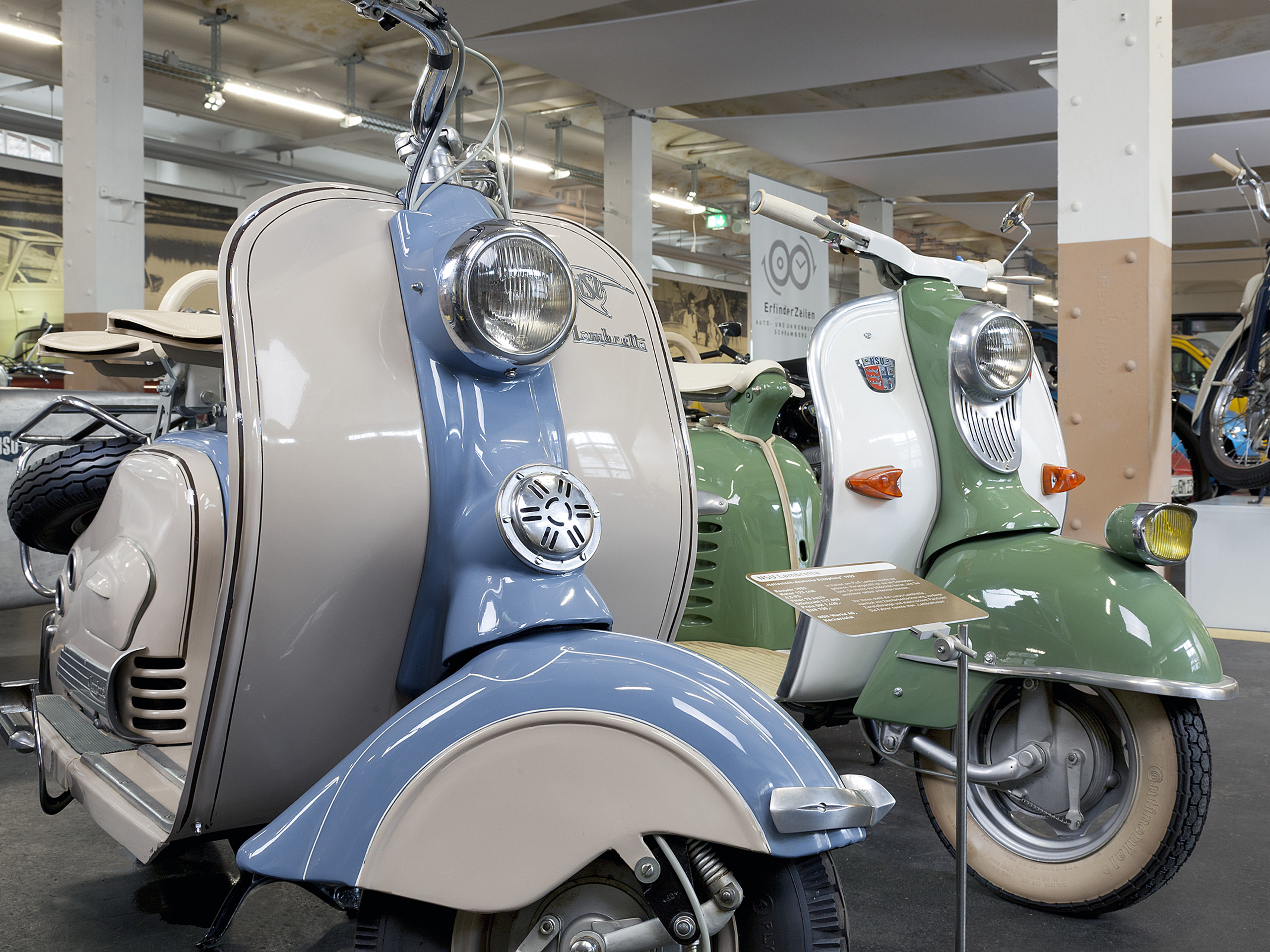 Zwei klassische Motorroller stehen in einem Ausstellungsraum. Der links ist in Beige und Blau gehalten, während der rechts grün und Weiß ist. Beide Roller haben runde Scheinwerfer und ansprechende Retro-Designs. Im Vordergrund ist ein Informationsschild sichtbar.