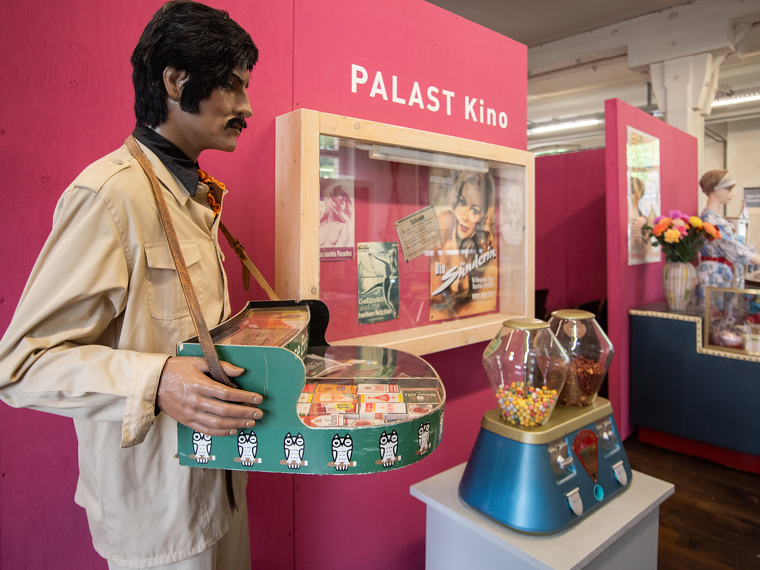 Ein Schaufenster mit einem schlichten, pinken Hintergrund zeigt eine Mannequin-Figur im beigen Anzug, die eine Verkaufsplatte mit Süßigkeiten hält. Im Hintergrund ist ein weiteres Display mit Filmplakaten und einem Blumenstrauß auf einem Tisch zu sehen. Oben steht "PALAST Kino".
